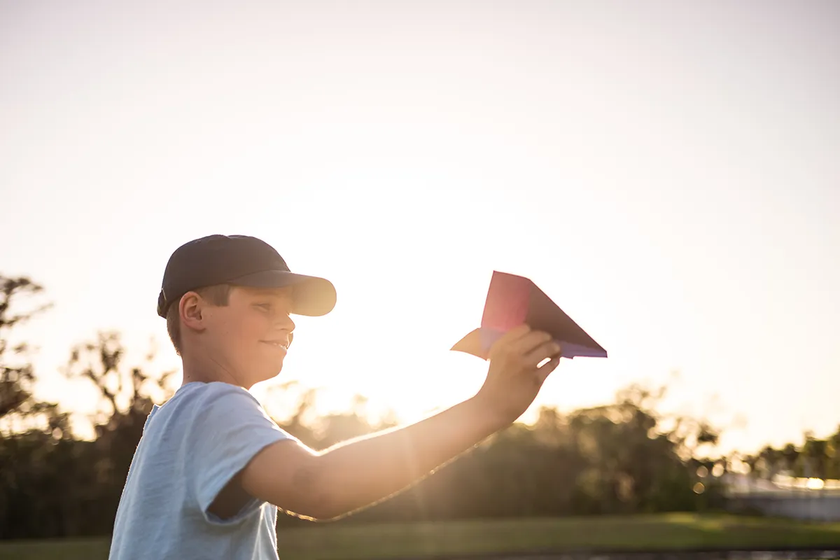 A boy wearing a cap smiles as he holds up a red paper airplane outdoors at sunset, with sunlight streaming behind him and trees in the background.