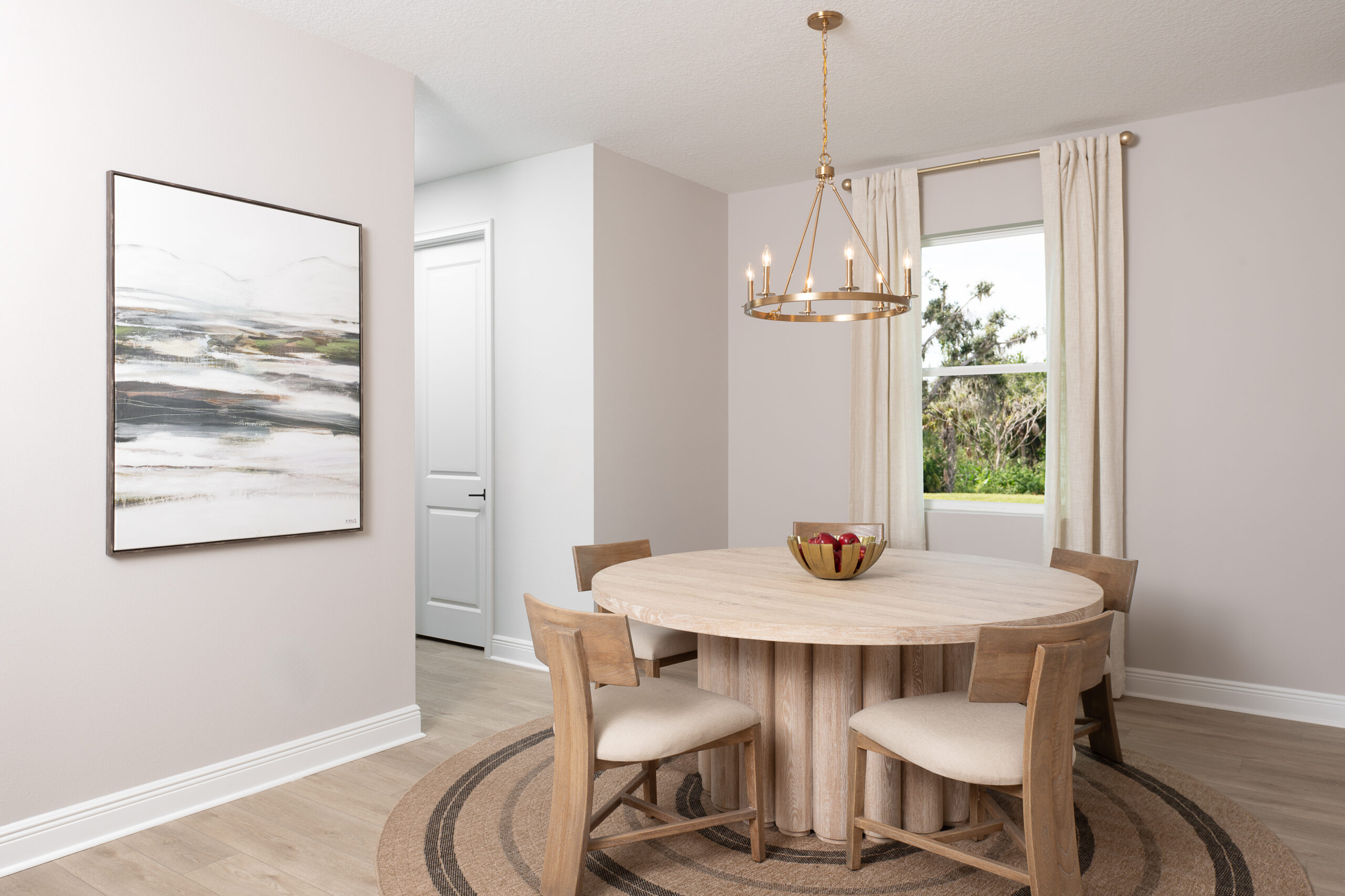 A modern dining room with a round wooden table, four cushioned chairs, a bowl of apples, a round rug, a window with curtains, a large abstract painting, and a gold chandelier.