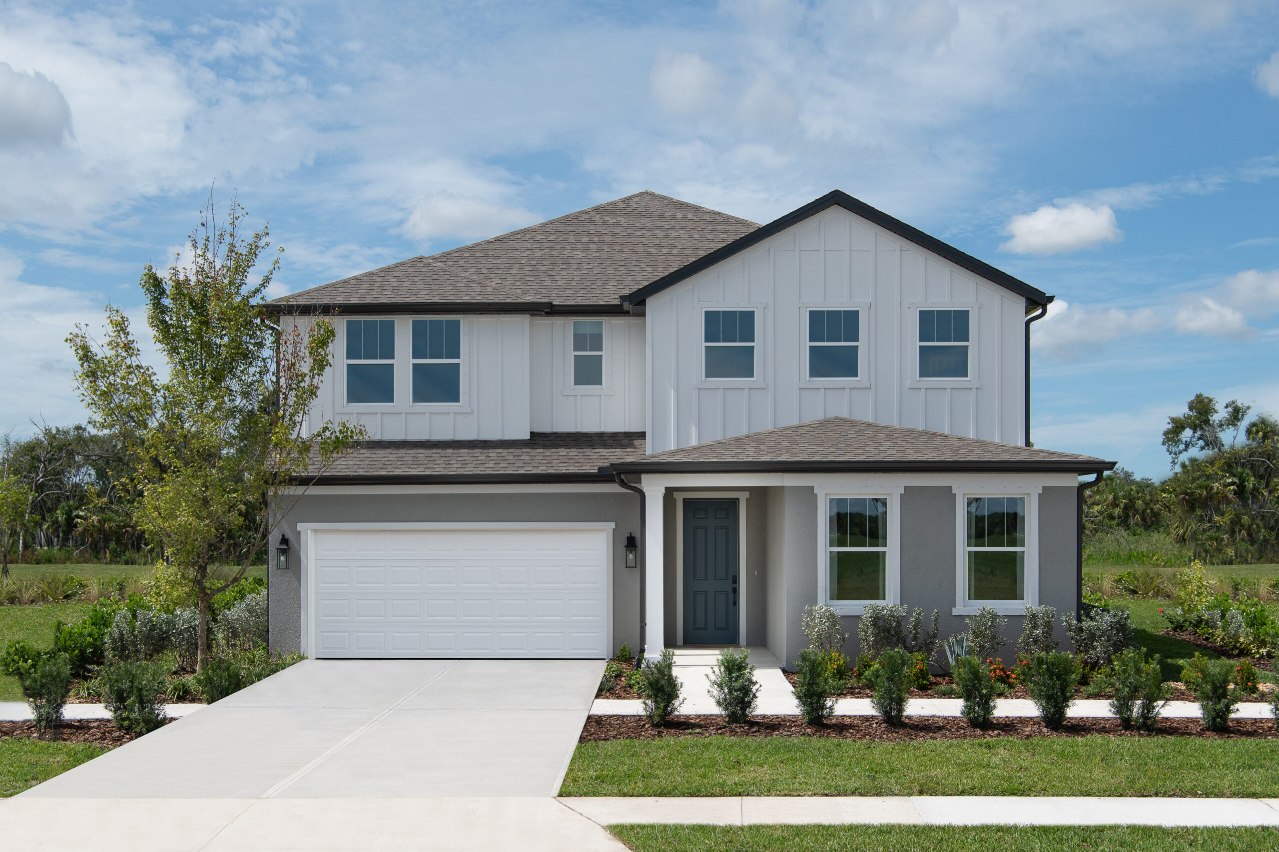 Two-story modern suburban house with a gray roof, white siding, and a two-car garage, surrounded by a neatly landscaped yard with shrubs and a small tree, set against a partly cloudy sky.