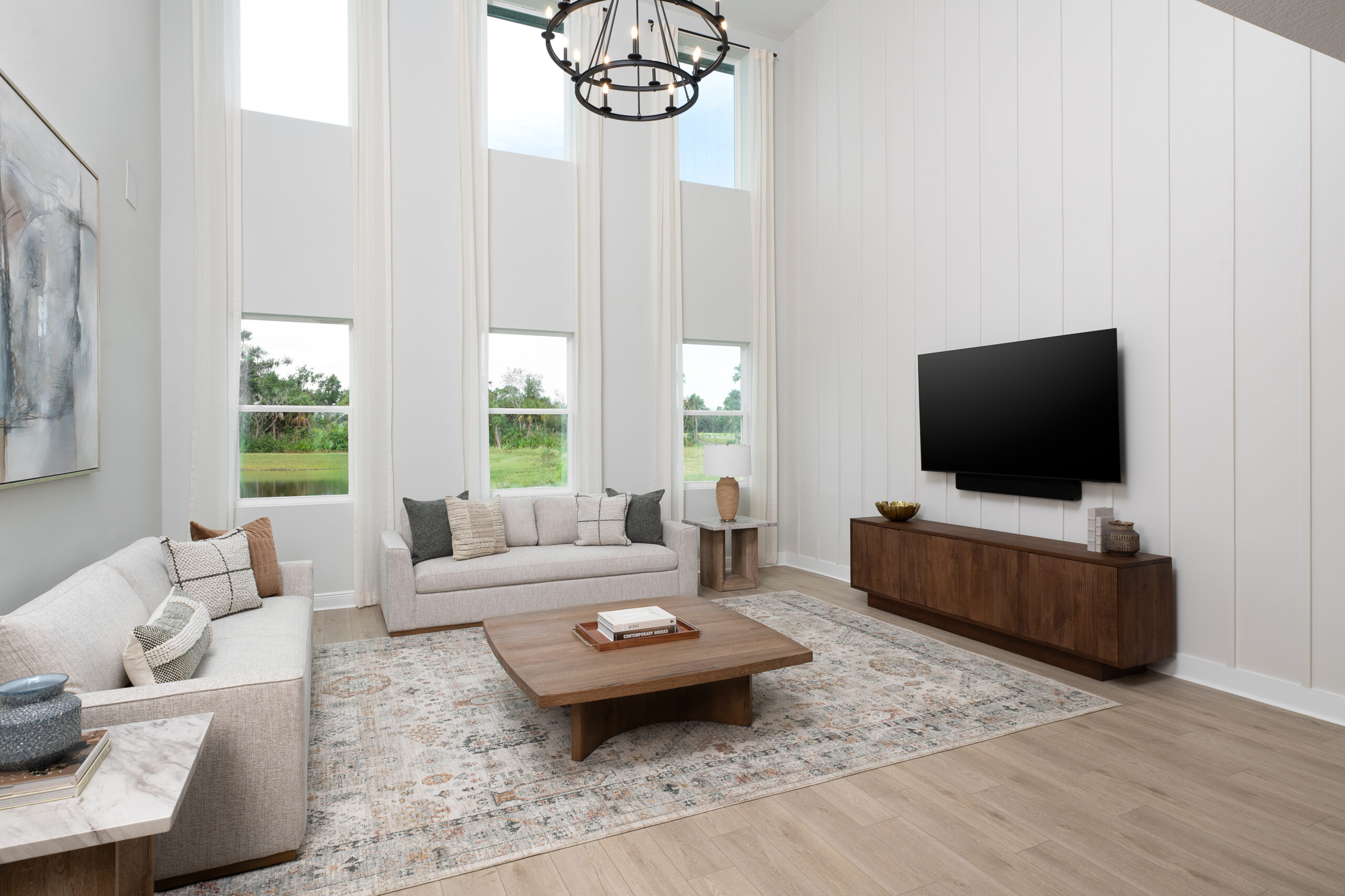 A modern living room with high ceilings, large windows, two beige sofas, a wooden coffee table, a wall-mounted TV on a wooden console, and light-colored decor with a patterned rug. Natural light fills the space.