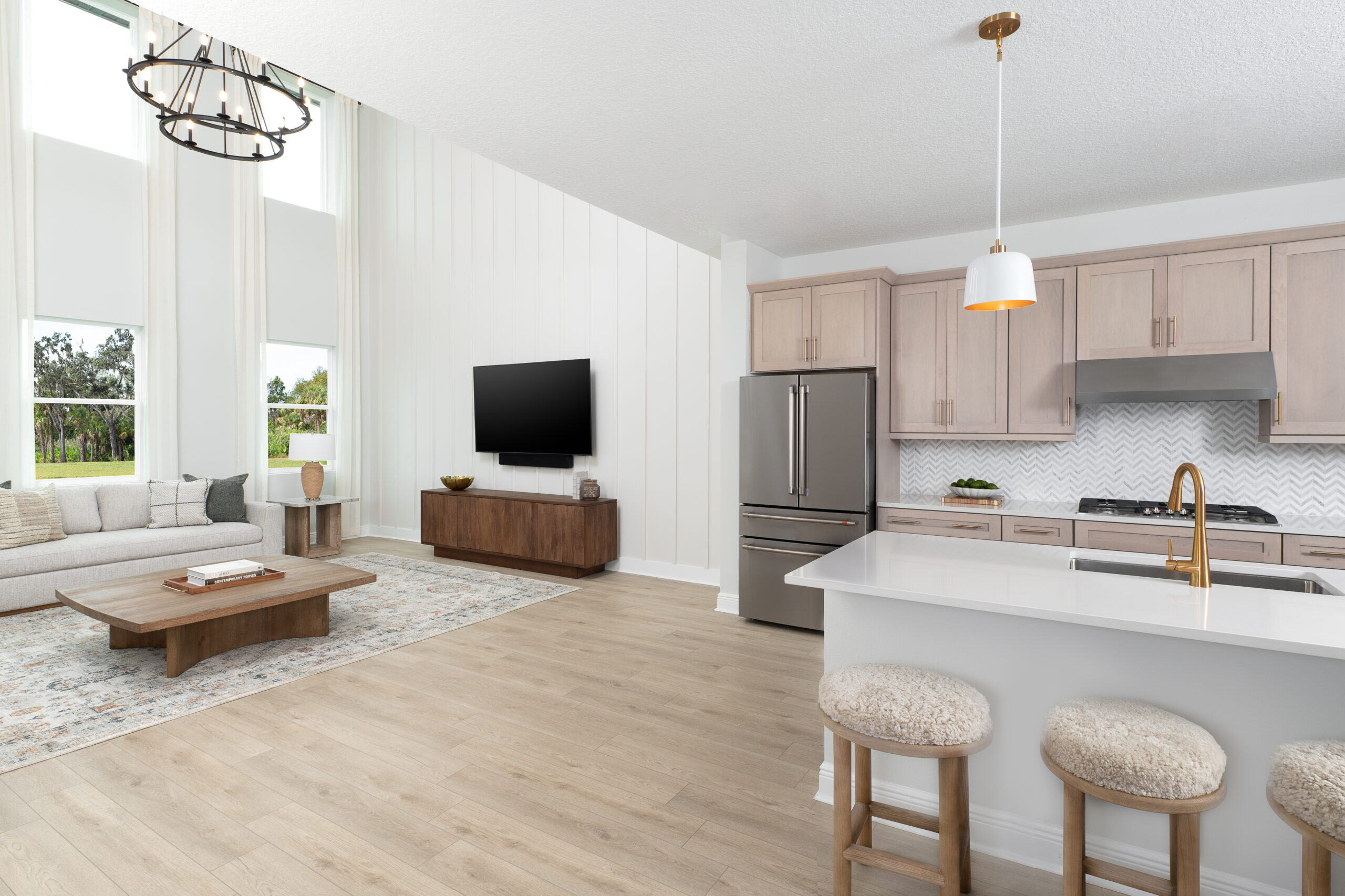 Modern open-concept living room and kitchen with light wood floors, beige cabinets, stainless steel appliances, a white island with two stools, a sofa, coffee table, wall-mounted TV, and large windows with natural light.