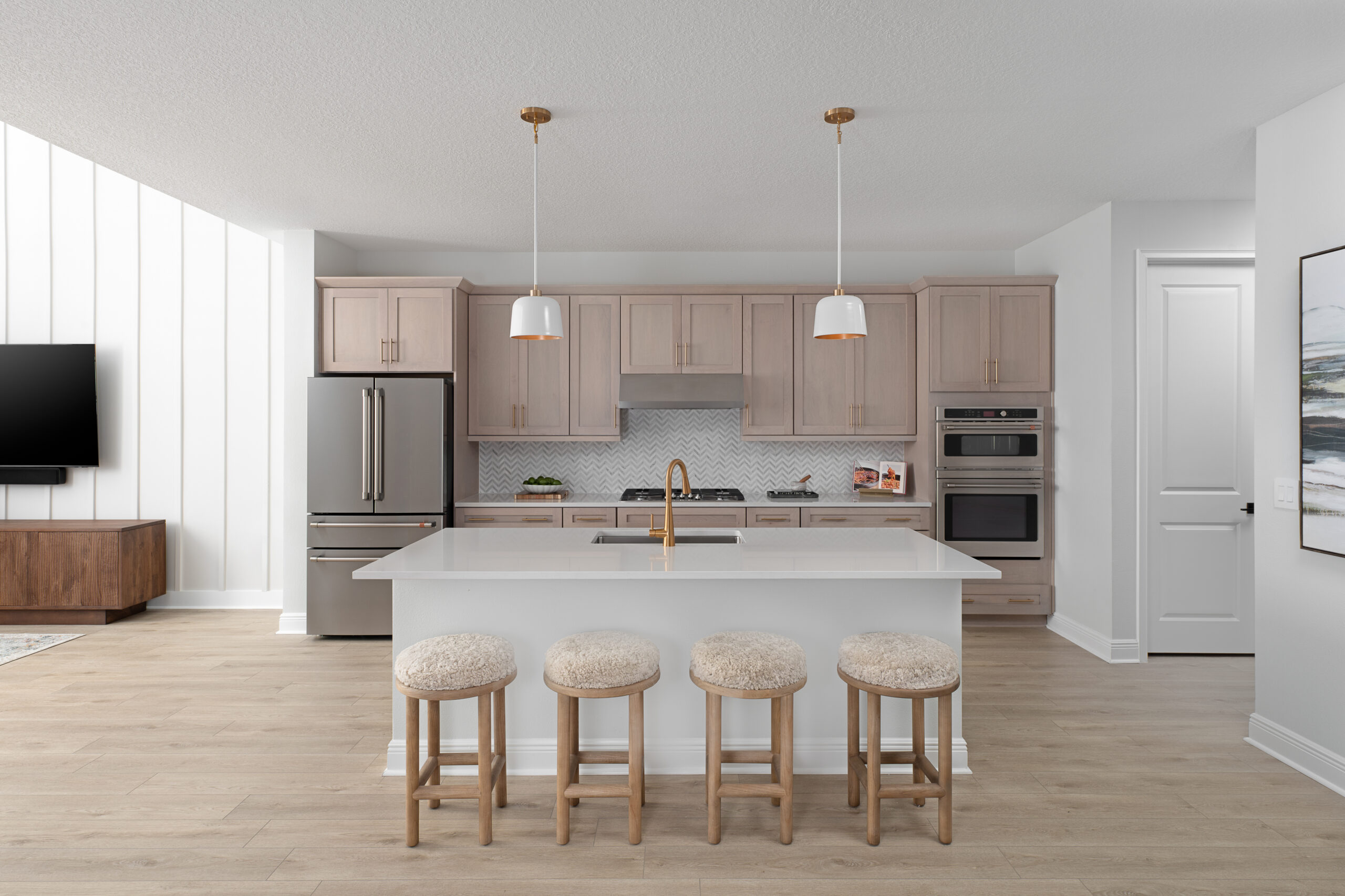 Modern kitchen with light wood cabinets, stainless steel appliances, a white island with a gold faucet, and four cushioned bar stools. Pendant lights hang above the island, and the space has light wood flooring.