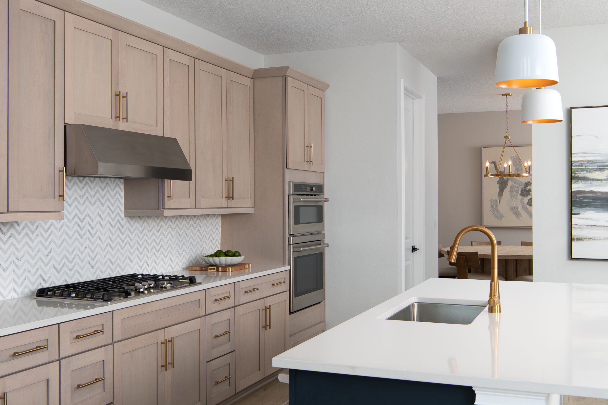 Modern kitchen with light wood cabinets, stainless steel appliances, a white herringbone backsplash, built-in oven, gas cooktop, white countertops, gold hardware and fixtures, and a glimpse of a dining area in the background.