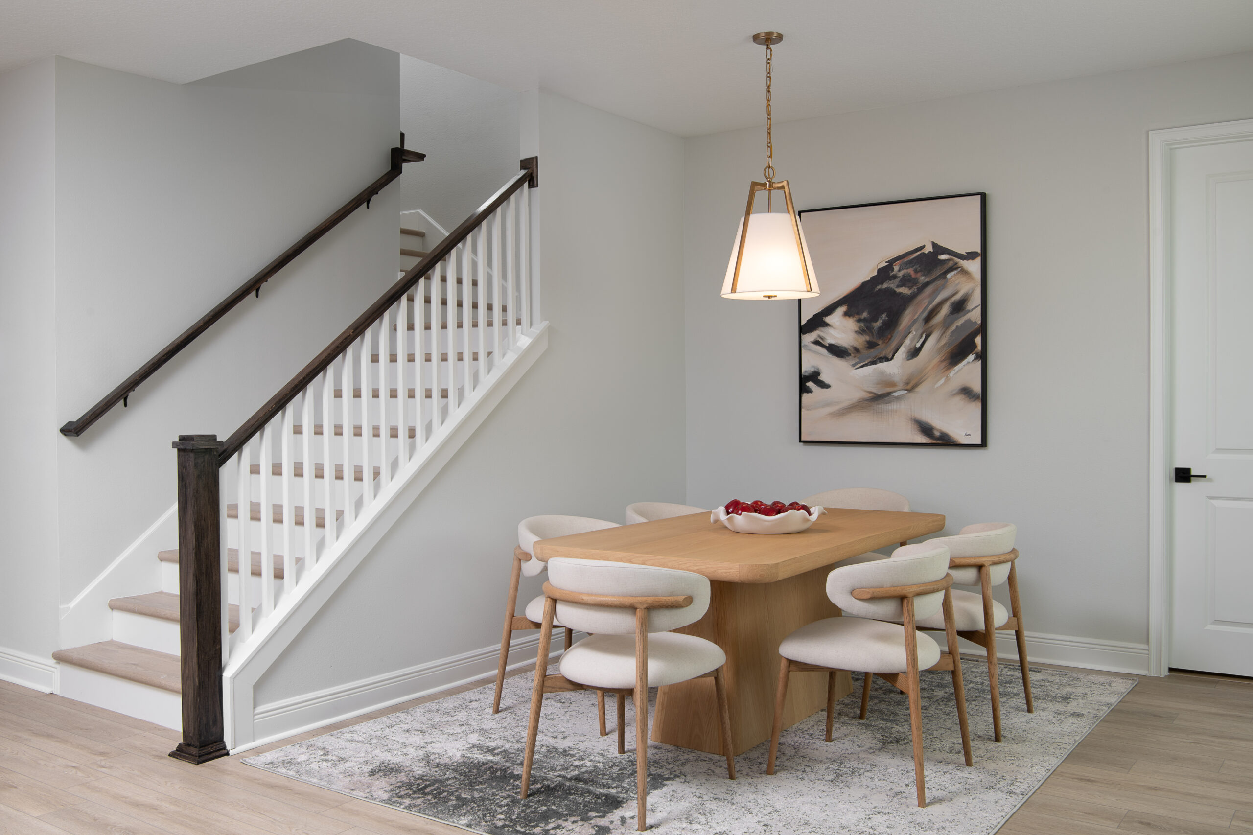 A modern dining area with a light wooden table and six cream-colored chairs sits on a patterned rug. A bowl of red apples is on the table. A staircase with dark railings and a mountain painting are in the background.