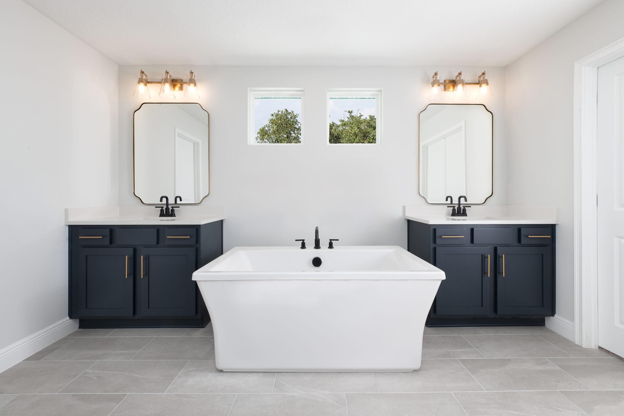 Modern bathroom with a white freestanding bathtub centered between two navy blue vanities, each with a mirror, sink, and wall-mounted lights. Two small windows above let in natural light.