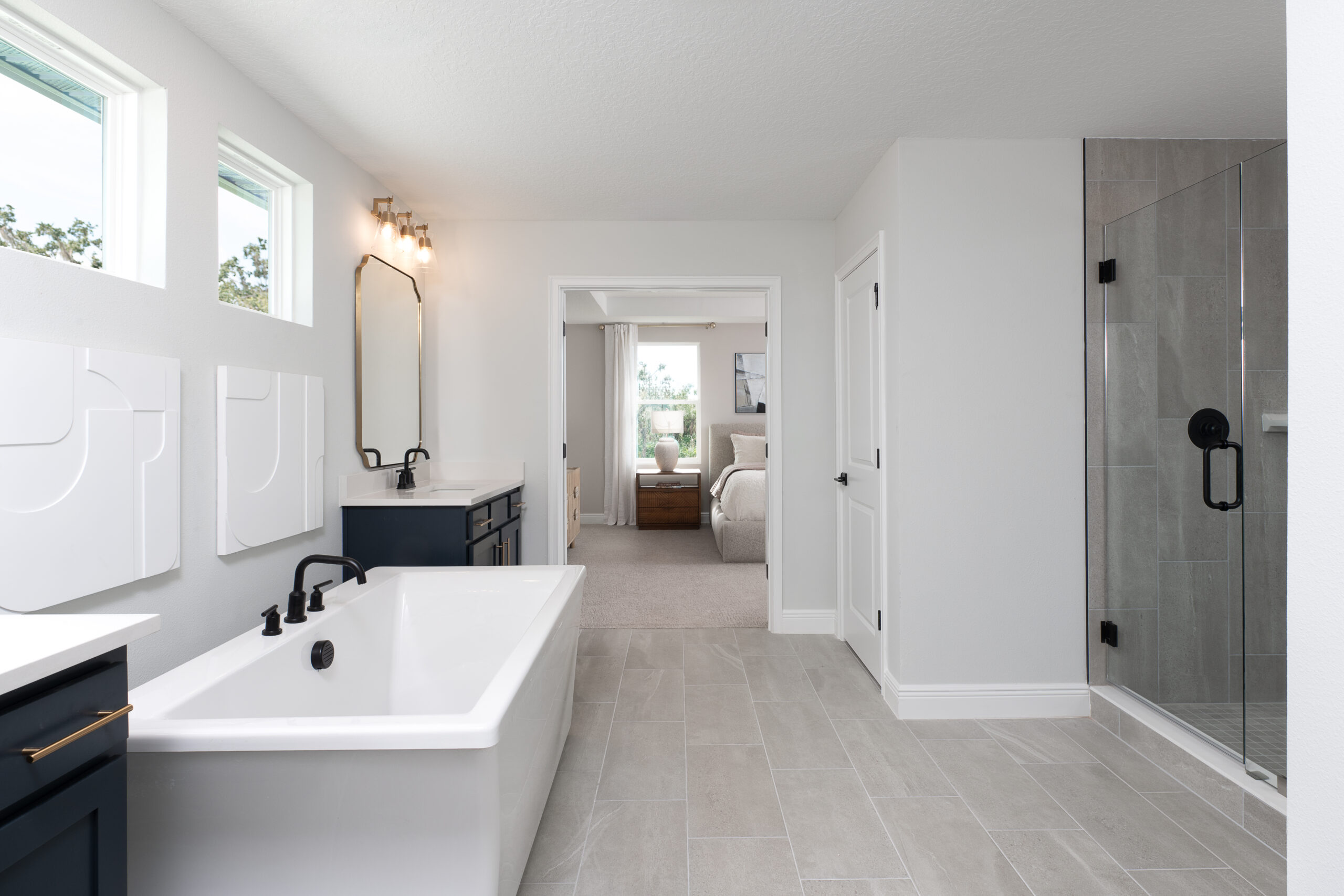 Modern bathroom with a soaking tub, black fixtures, dual vanity, large wall mirror, glass-enclosed shower, light gray tile floor, and an open doorway leading to a cozy bedroom.