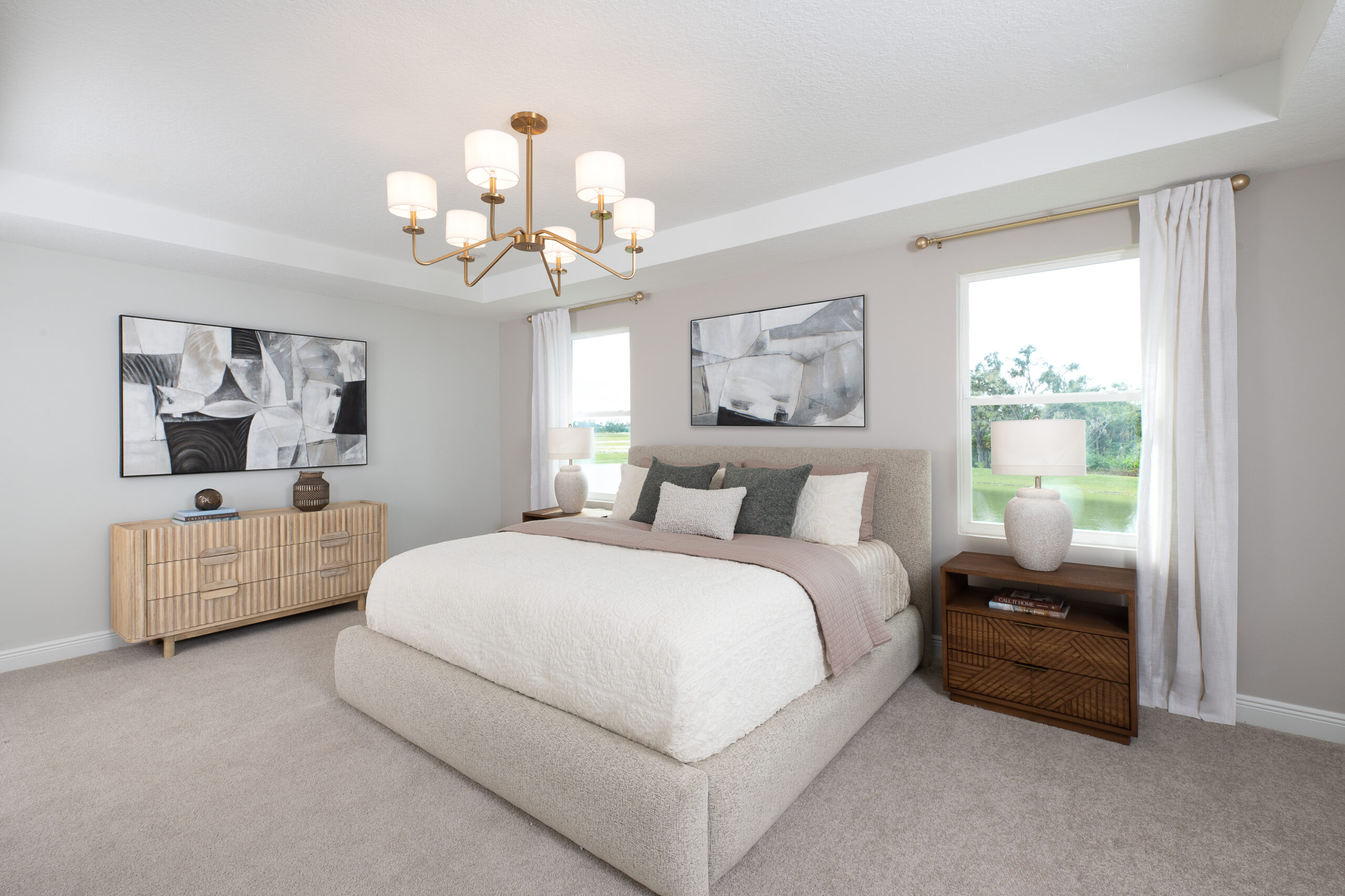 Modern bedroom with a large bed, beige carpet, two nightstands with lamps, a dresser, abstract wall art, and a large window with white curtains letting in natural light.