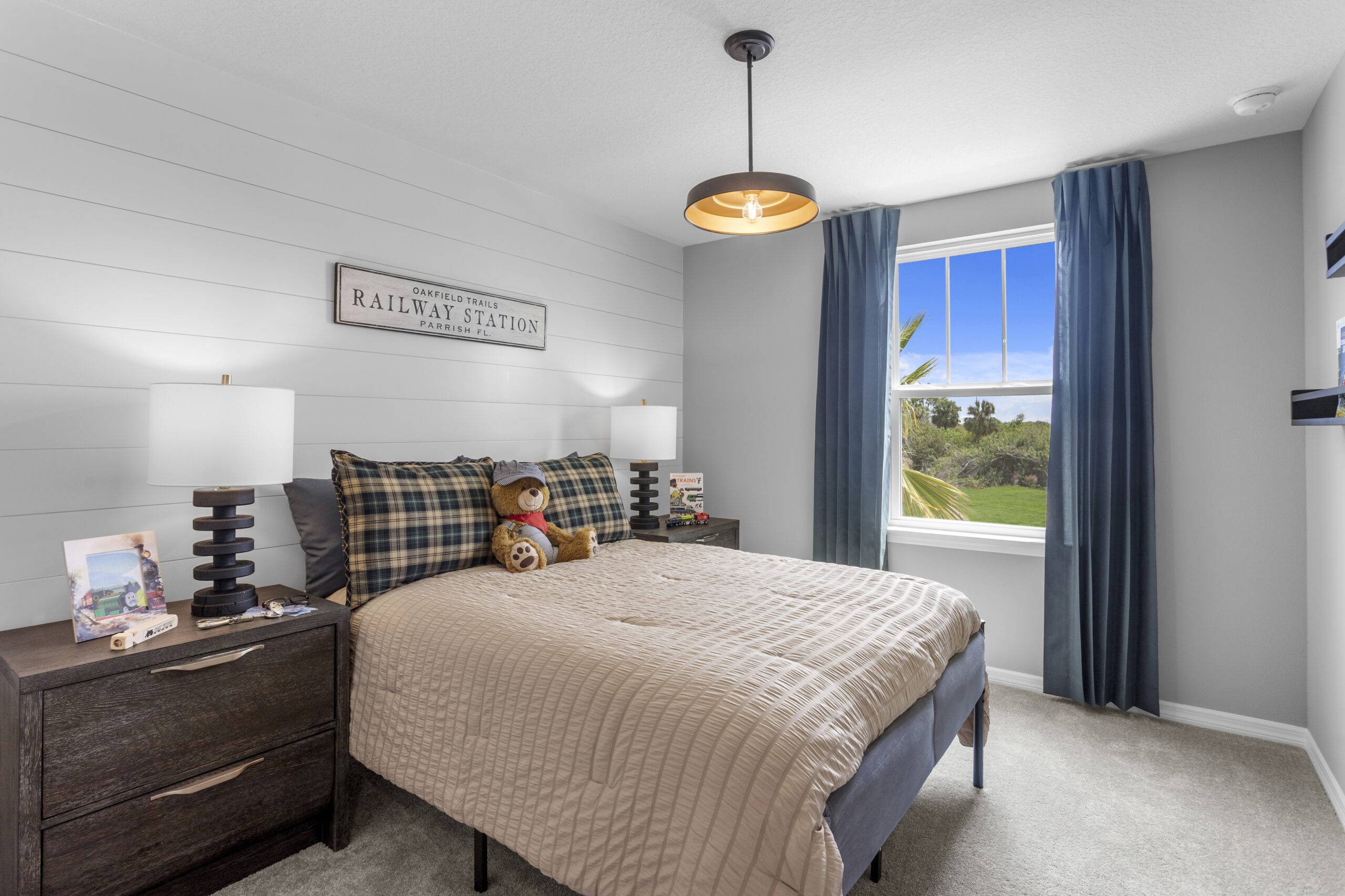 A cozy bedroom with a bed, plaid pillows, and a teddy bear. Two nightstands hold lamps and books. A window with blue curtains lets in sunlight, and a “Railway Stations” sign decorates the shiplap wall.
