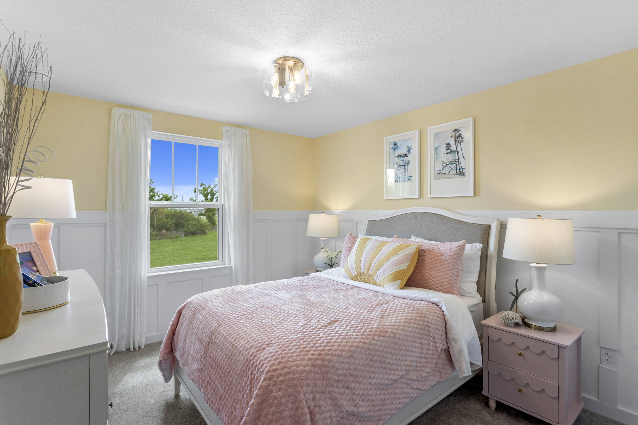A cozy bedroom with light yellow and white walls, a bed with a pink textured comforter, two bedside tables with lamps, a dresser, a window with white curtains, and framed art above the bed.