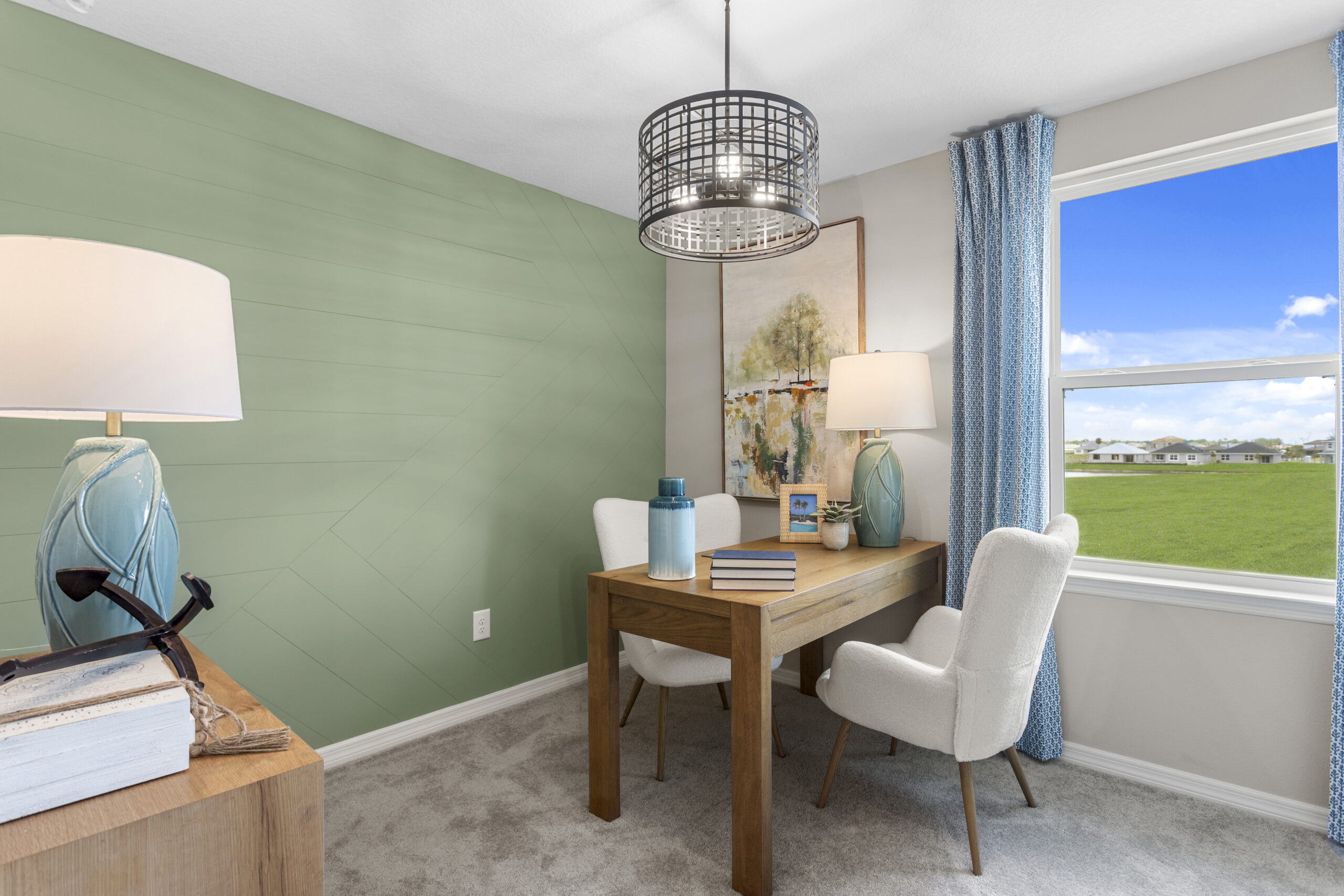 A bright home office with a wooden desk, two white chairs, blue lamps, books, artwork, and a large window showing a green field and blue sky. Light green accent wall and modern ceiling light fixture.