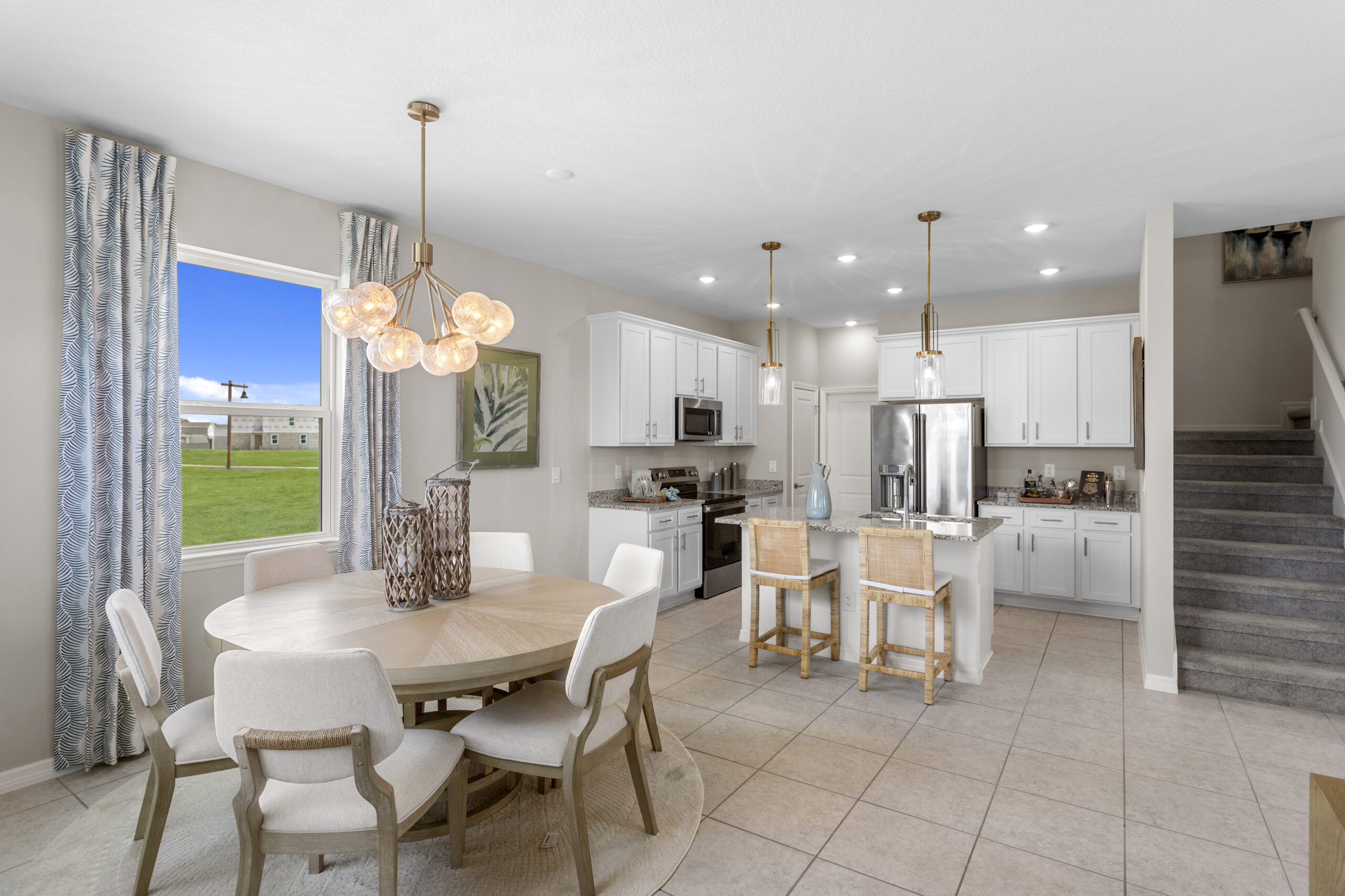 Bright, modern kitchen and dining area with white cabinets, stainless steel appliances, light wood furniture, pendant lighting, and large window overlooking green lawn; carpeted stairs on the right.