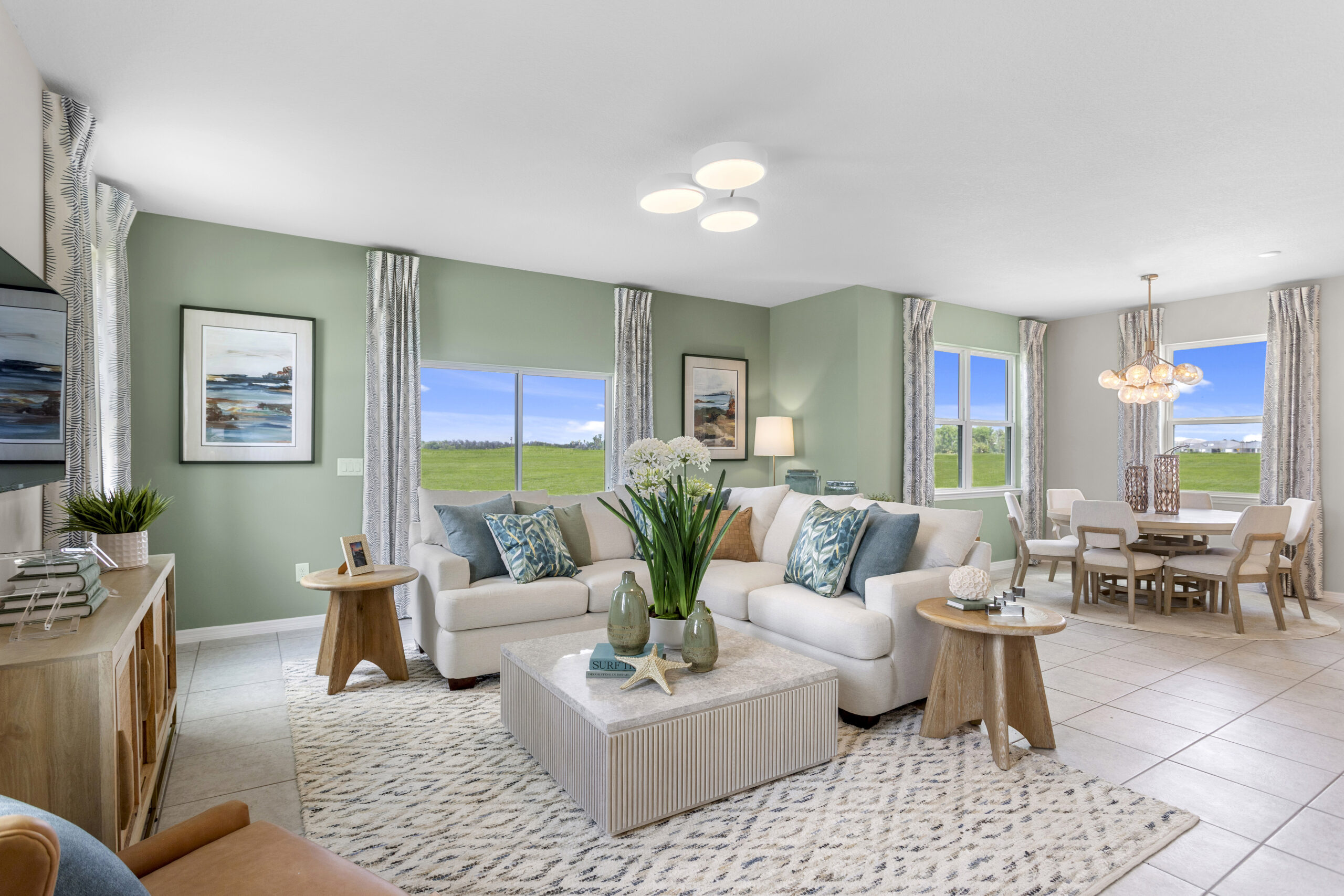 Bright, modern living and dining area with light-colored furniture, decorative pillows, a patterned rug, large windows showing green landscape, and contemporary decor elements. Dining table and chairs are visible in the background.