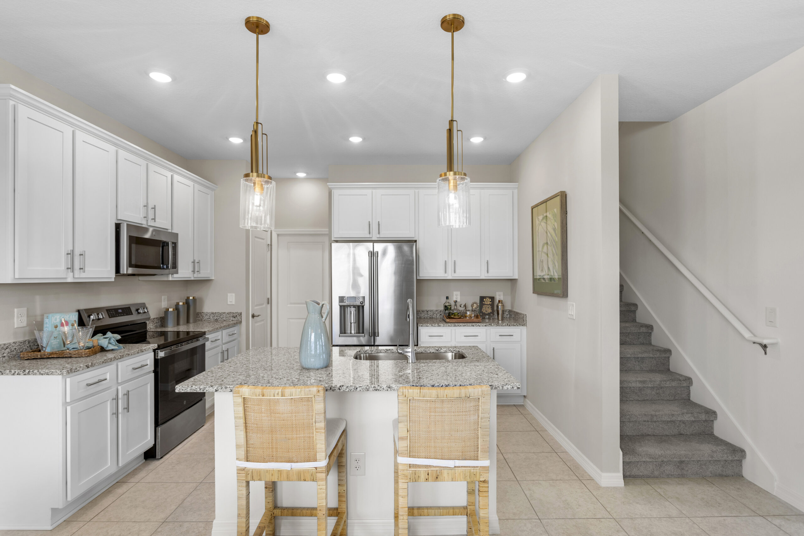 Modern kitchen with white cabinets, stainless steel appliances, granite countertops, an island with two wicker bar stools, pendant lights above, and a staircase with carpeted steps on the right.