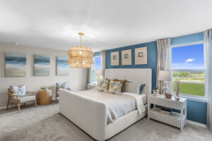 Bright, modern bedroom with a large bed, blue accent wall, three abstract art pieces, two windows with outdoor views, wicker pendant light, two nightstands, and cozy seating area with chairs and pillows.