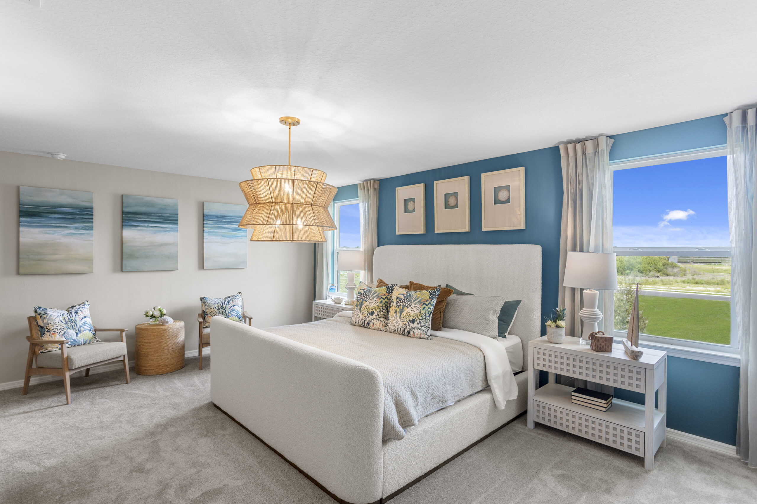 Bright, modern bedroom with a large bed, blue accent wall, three abstract art pieces, two windows with outdoor views, wicker pendant light, two nightstands, and cozy seating area with chairs and pillows.