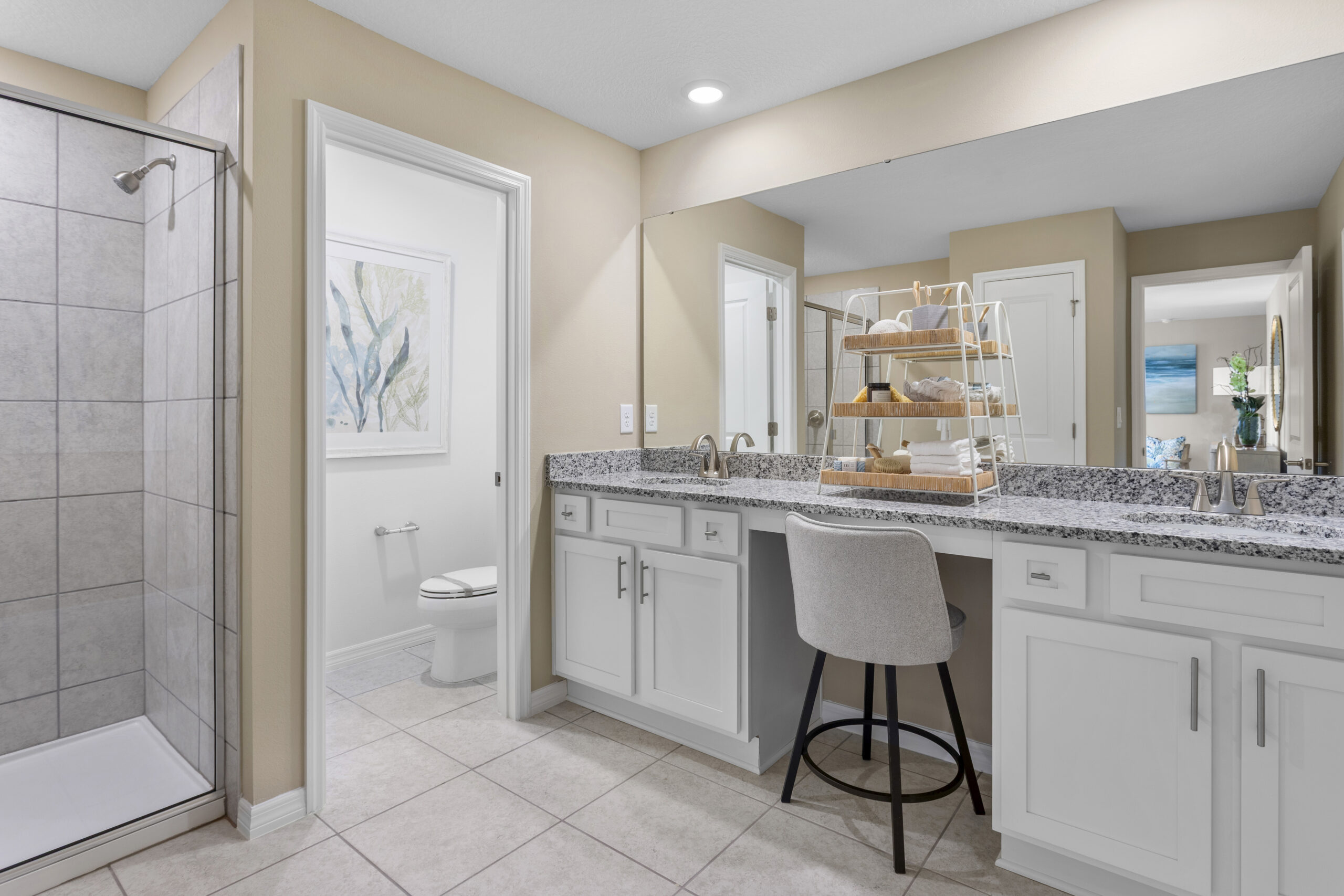 Modern bathroom with a granite countertop, white cabinets, a built-in vanity with a cushioned stool, a walk-in shower with tiled walls, and a toilet in a separate small room. Large mirror reflects hallway decor.