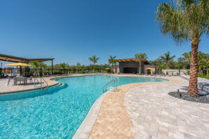 A large outdoor swimming pool with clear blue water, surrounded by palm trees and a paved deck area with lounge chairs and tables under yellow umbrellas, on a sunny day.