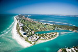 Aerial view of a narrow strip of land with white sandy beaches, turquoise water, resorts, and residential buildings, bordered by ocean on both sides under a clear blue sky.
