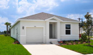 A single-story white house with a gray roof, attached garage, front porch, and small landscaped garden, set against a blue sky with scattered clouds and surrounded by green grass.