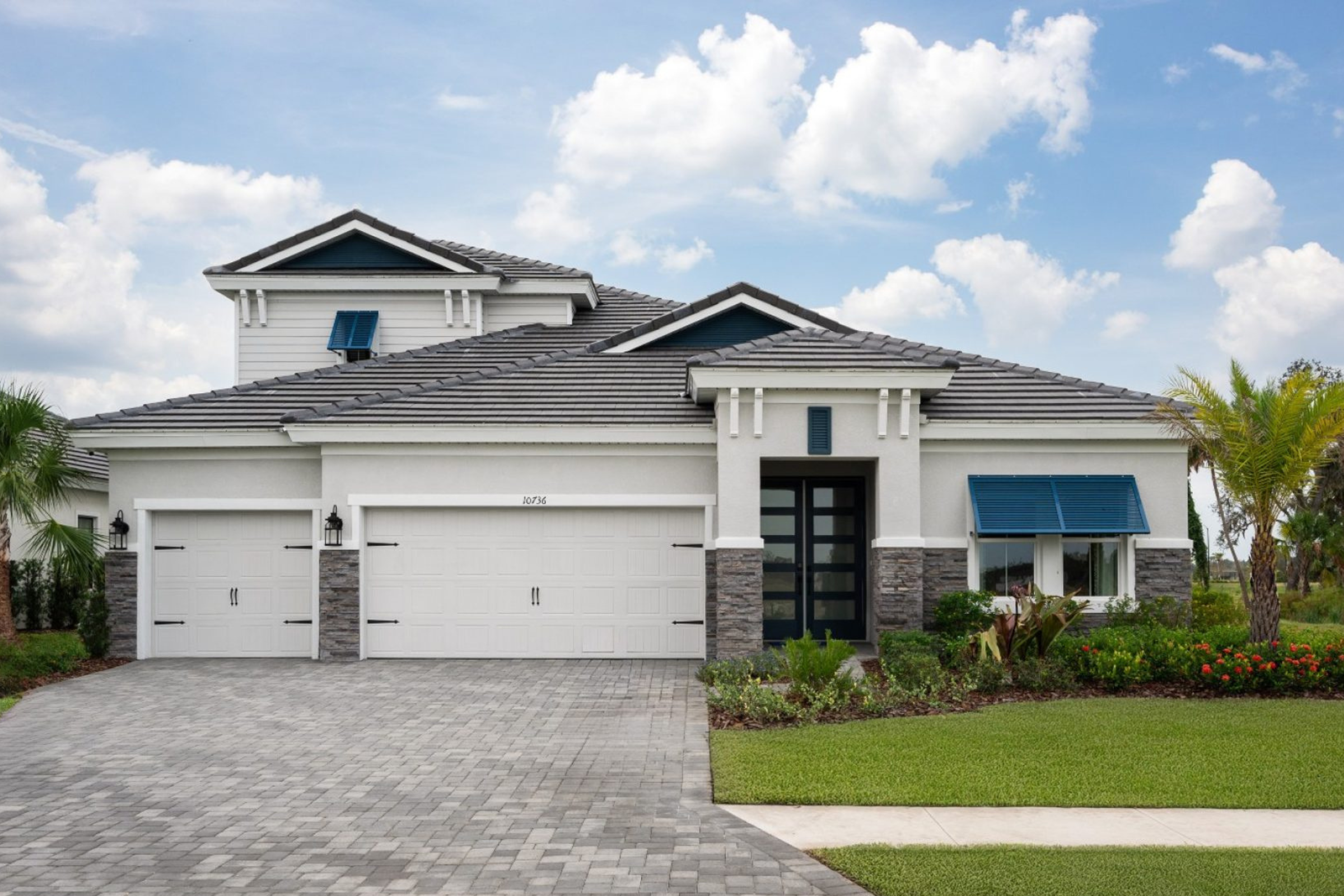 Modern two-story house with a gray tiled roof, white and stone siding, three garage doors, blue window awnings, a paved driveway, and a well-maintained lawn with palm trees and shrubs under a partly cloudy sky.