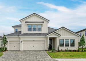 Modern two-story house with light-colored siding, a three-car garage, multiple large windows, a gray tiled roof, a paved driveway, and landscaped front yard, set under a blue sky.