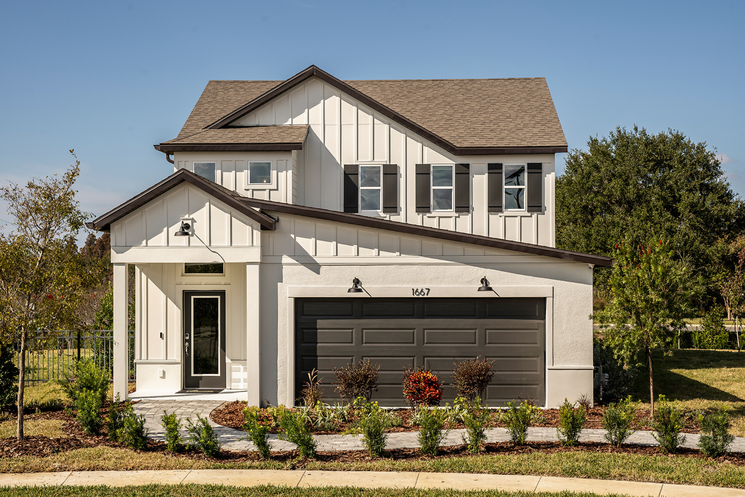 Two-story modern suburban house with white siding, dark trim, and a double garage. The front yard features neatly landscaped plants and a small pathway, and the house number 1667 is visible above the garage.