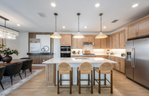 Modern kitchen with light wood cabinets, stainless steel appliances, a large island with three bar stools, and pendant lights. An adjacent dining area features a dark table, black chairs, and a chandelier.