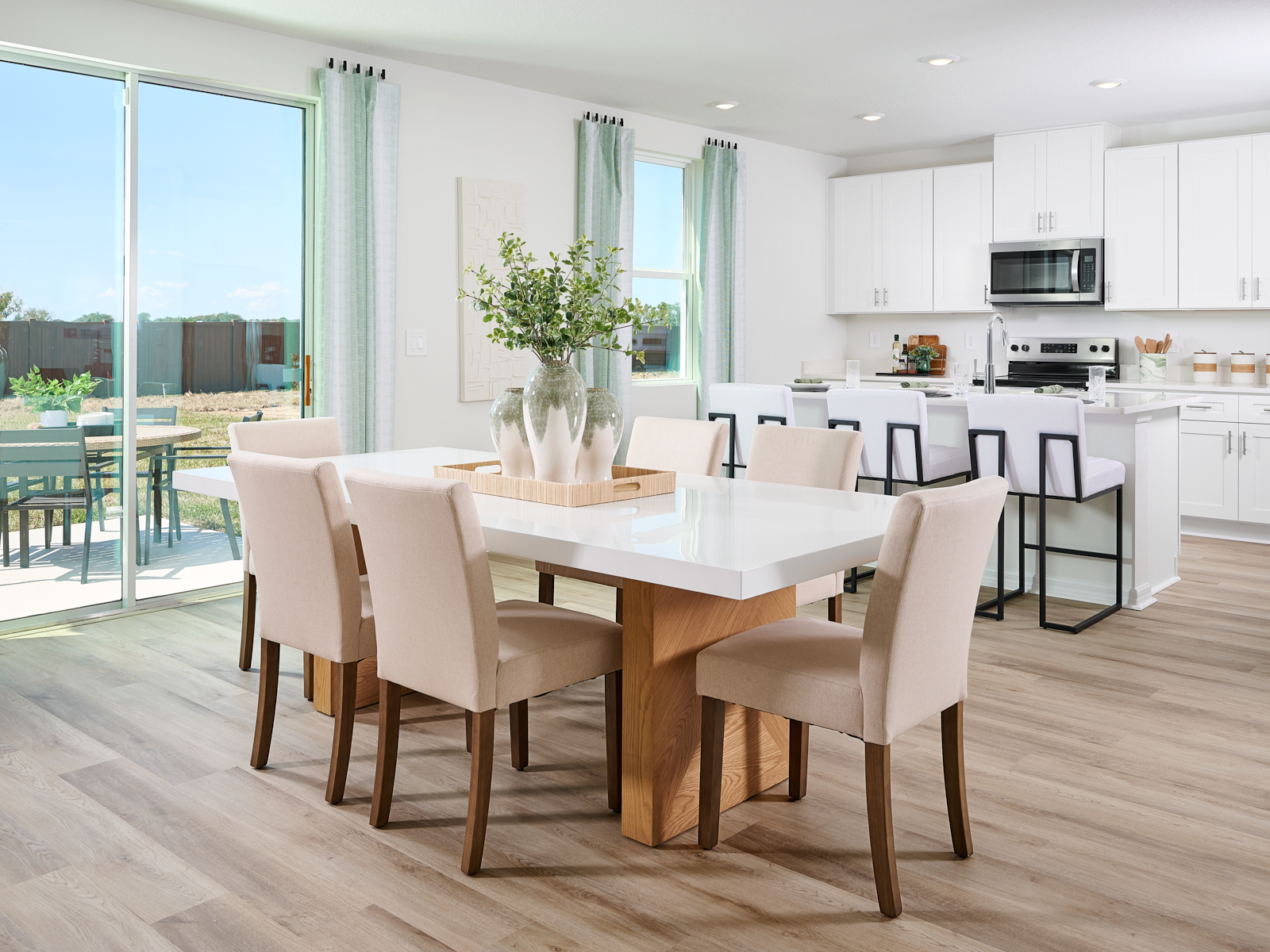 Bright, modern dining and kitchen area with a white table, beige chairs, wood floors, white cabinets, and large windows showing a sunny backyard with a patio table and chairs.