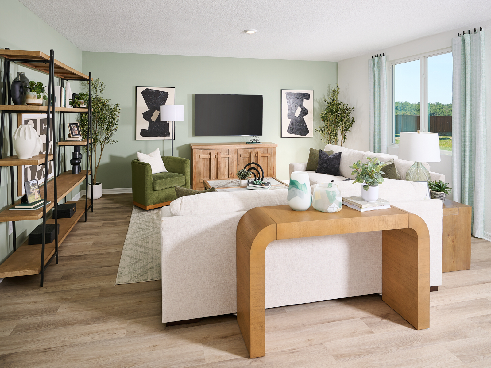 A modern living room with light wood floors, a beige sofa, green armchair, wooden console, TV, bookshelf, two abstract art pieces, potted plants, and large windows with sheer curtains letting in natural light.