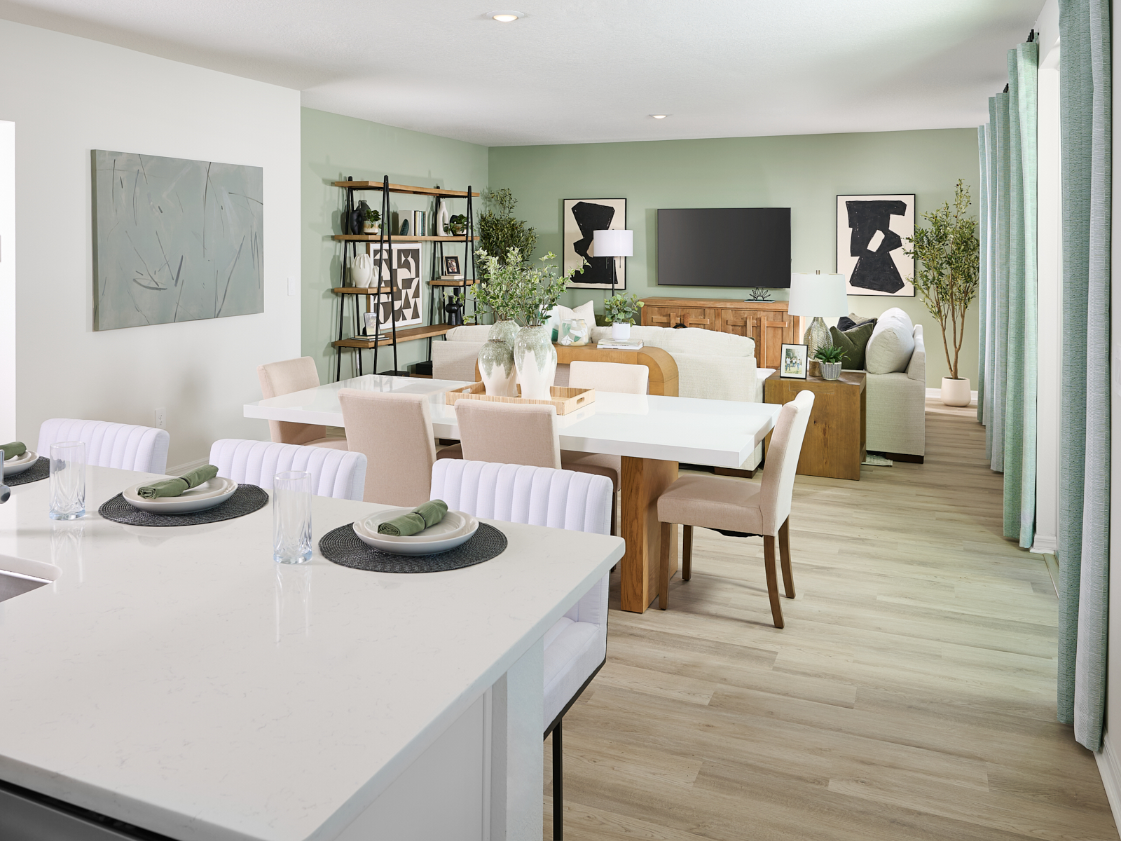 Modern open-plan dining and living area with light green walls, large windows with curtains, a white dining table set for four, neutral-toned furniture, wall art, shelves with plants, and a TV on a wooden console.