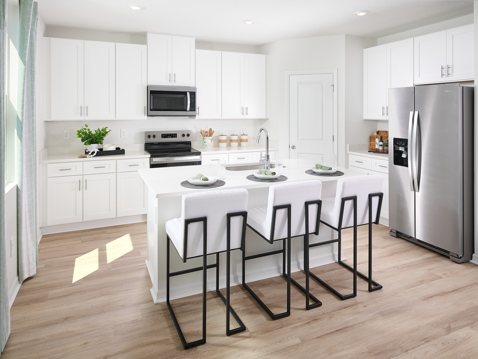 Modern white kitchen with stainless steel appliances, an island with a sink, and three white bar stools with black metal frames. Sunlight streams in from a window, illuminating the wood floor and minimalist decor.
