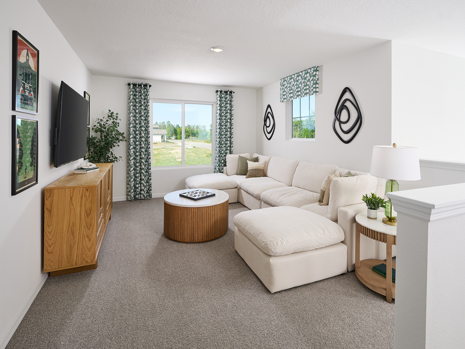 A bright living room with a large white sectional sofa, round wooden coffee table, TV on a wooden stand, framed art, patterned curtains, abstract wall art, and a window with a view of greenery outside.