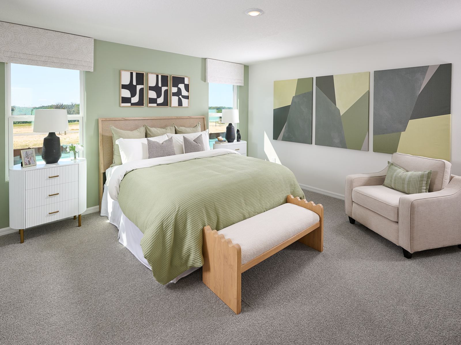 A modern bedroom with a bed featuring green and white bedding, two white nightstands with lamps, abstract art on the wall, a beige armchair, and a wooden bench at the foot of the bed. Sunlight comes in through two windows.