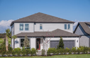 A modern two-story house with light gray siding, dark gray roof, a double garage, and a landscaped front yard with small trees and shrubs under a clear blue sky.